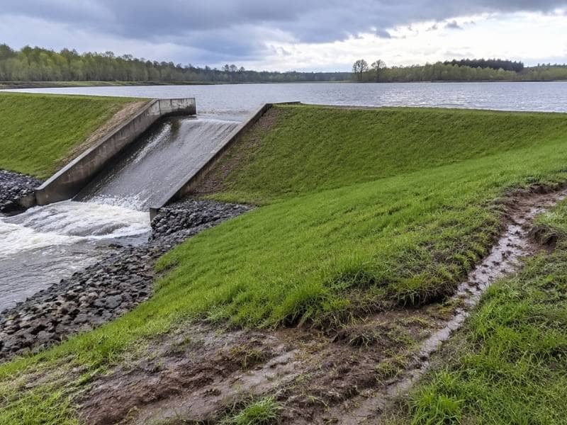 A dam inspection after heavy rainfall shows water flowing through the spillway and erosion along the embankment
